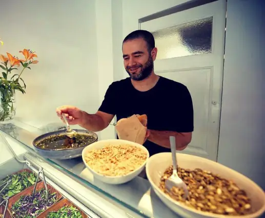 En mand, der ser ud til at være ejeren, står bag en madvogn og betjener en lækker portion mad. Han er i færd med at hælde en grøn sauce over maden og holder et stykke fladbrød i den anden hånd. Han smiler mens han forbereder maden, hvilket tilføjer en personlig og venlig atmosfære til oplevelsen. Foran ham er der skåle med forskellige tilbehør, herunder en skål med nødder og en anden med syltede grøntsager, alt sammen præsenteret i en appetitvækkende og indbydende måde