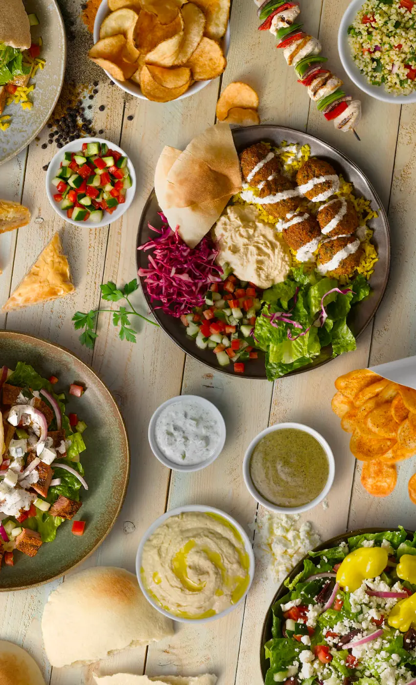 Et lyst træbord dækket med forskellige retter fra et middelhavsinspireret måltid. I midten af bordet er der en stor tallerken med gyldenbrune falafelkugler på en seng af gult krydret ris, garneret med tahinidressing og ledsaget af en klump cremet hummus og frisk rødkålssalat. Til venstre for denne tallerken er der en skål med en grøn salat toppet med store stykker af gyldenbrunt brød og fetaost, og en lille skål med en frisk agurk- og tomat-salat. I forgrunden er der et par pitabrød, halvt brækket. Til højre ses en spydpind med grillet kød og grøntsager og en skål med bulgursalat. Der er også forskellige skåle med dips, såsom en med en tyk hvid tzatzikisauce, en med en grøn krydret sauce og en med en mere flydende, klar dressing. En anden skål viser sprøde kartoffelchips, som er arrangeret stående. Hele arrangementet skaber en indbydende atmosfære og indikerer en afbalanceret blanding af farverige, friske og nærende elementer.