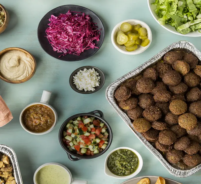 Et bord dækket med et udvalg af madvarer, der udgør en cateringpakke. I midten ses en stor aluminiumsform fyldt med gyldenbrune falafelkugler. Til højre for den er der en skål med sprøde kartoffelchips. Øverst på billedet er der et træfad med friske æbler, en bakke med baklava skåret i trekanter, og en skål med store, hjemmebagte cookies. Forskellige skåle indeholder friskhakket salat, rødkålssalat, syltede agurker og en lille skål med hvidløg. Der er også små skåle med forskellige saucer og dips, herunder hummus, salsa og grønne samt hvide cremede saucer. En kurv med bløde pitabrød og en tallerken med en anden form for bagværk ligger også på bordet. Forgrunden viser en orange tallerken med sølvbestik samt to lyserøde drikke med citronskiver. Hele scenen udstråler en hyggelig frokost eller middag til en større gruppe, arrangeret på en lys turkis baggrund, der giver et frisk og indbydende look.