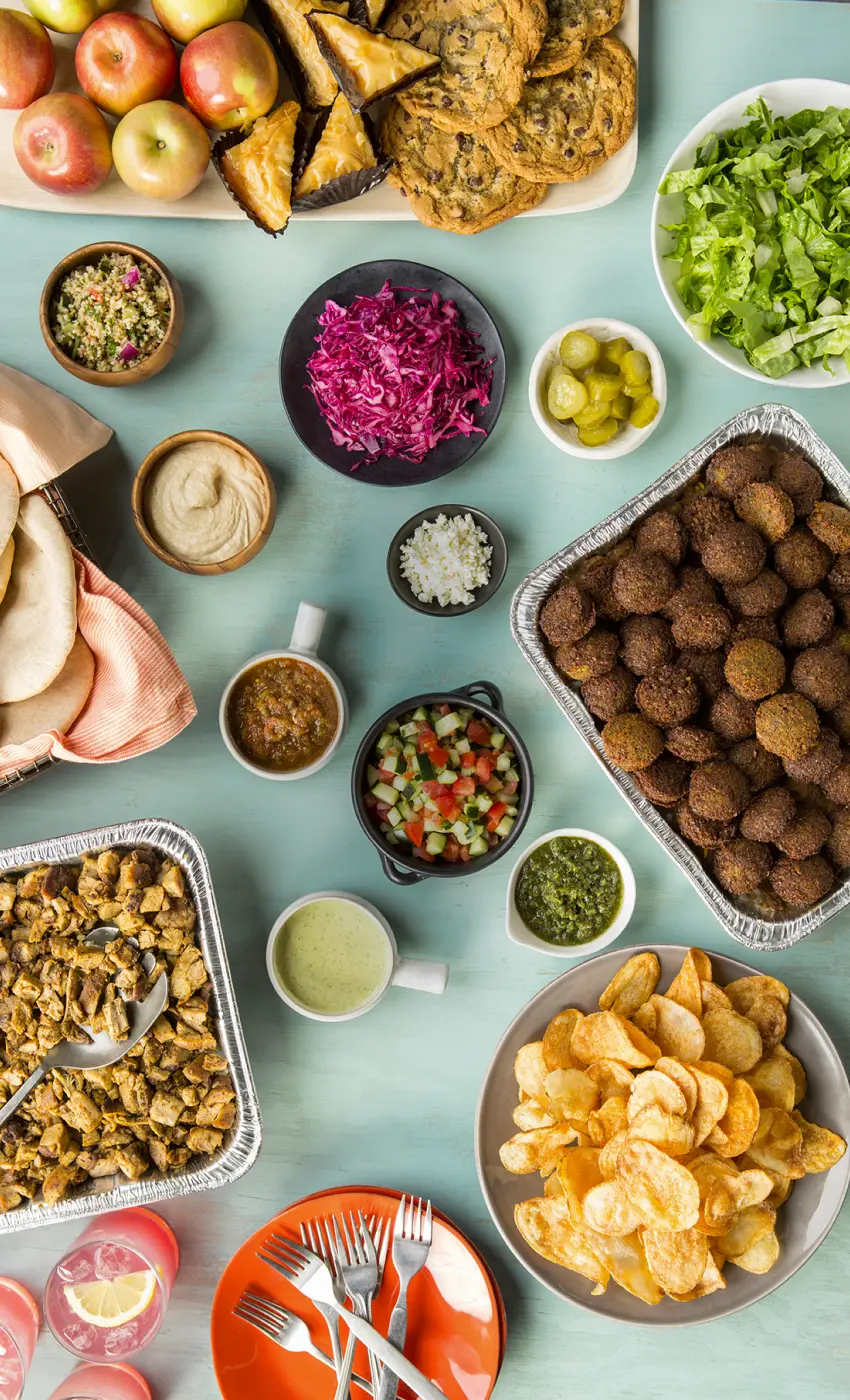 Et bord dækket med et udvalg af madvarer, der udgør en cateringpakke. I midten ses en stor aluminiumsform fyldt med gyldenbrune falafelkugler. Til højre for den er der en skål med sprøde kartoffelchips. Øverst på billedet er der et træfad med friske æbler, en bakke med baklava skåret i trekanter, og en skål med store, hjemmebagte cookies. Forskellige skåle indeholder friskhakket salat, rødkålssalat, syltede agurker og en lille skål med hvidløg. Der er også små skåle med forskellige saucer og dips, herunder hummus, salsa og grønne samt hvide cremede saucer. En kurv med bløde pitabrød og en tallerken med en anden form for bagværk ligger også på bordet. Forgrunden viser en orange tallerken med sølvbestik samt to lyserøde drikke med citronskiver. Hele scenen udstråler en hyggelig frokost eller middag til en større gruppe, arrangeret på en lys turkis baggrund, der giver et frisk og indbydende look